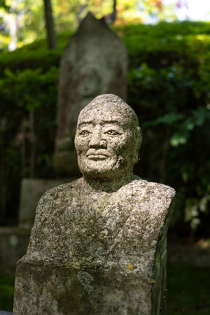 A weathered stone statue of a serene figure stands in quiet contemplation at Mitaki-Dera Temple in Hiroshima, surrounded by forest shadeの写真素材