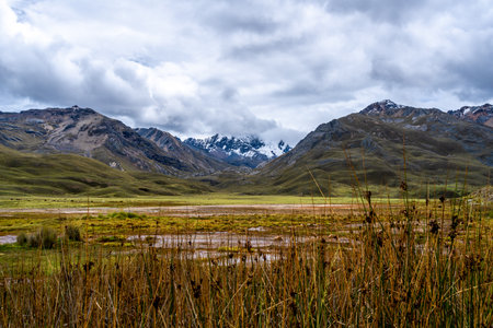 A marshy lake reflecting the dramatic cloudy sky is surrounded by rolling hills and snow-capped...