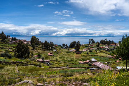 Traditional houses with red roofs and cultivated fields are spread across the green landscape of Taquile Island, overlooking the deep blue waters of Lake Titicaca in Peru. A peaceful rural settingの写真素材