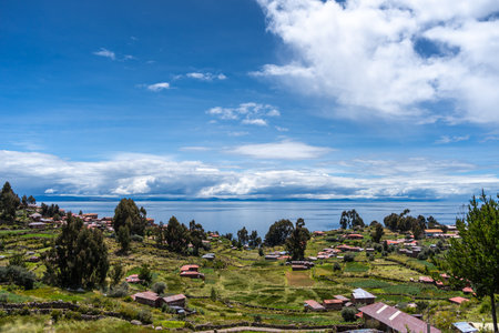 Traditional houses with red roofs and cultivated fields are spread across the green landscape of Taquile Island, overlooking the deep blue waters of Lake Titicaca in Peru. A peaceful rural settingの写真素材