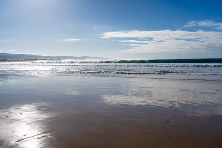 A vast sandy beach stretches along the Great Ocean Road, Australia, with gentle waves lapping the shore under a partly cloudy skyの写真素材