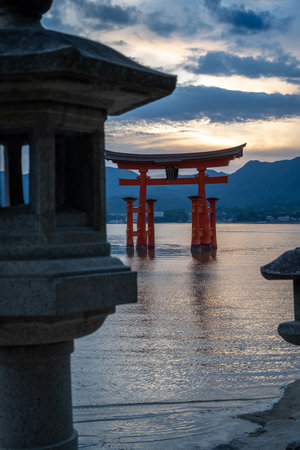 The famous floating torii gate of Itsukushima Shrine stands in calm waters at sunset, framed by stone lanterns and distant mountainsの写真素材
