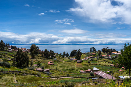 Traditional houses with red roofs and cultivated fields are spread across the green landscape of Taquile Island, overlooking the deep blue waters of Lake Titicaca in Peru. A peaceful rural settingの写真素材