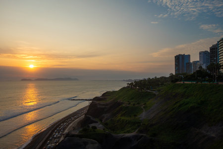 The sun sets over Lima, Peru, casting a golden glow on the Pacific Ocean and the green cliffs. The serene coastal scenery offers a breathtaking view of natureâs beauty and tranquilityの写真素材