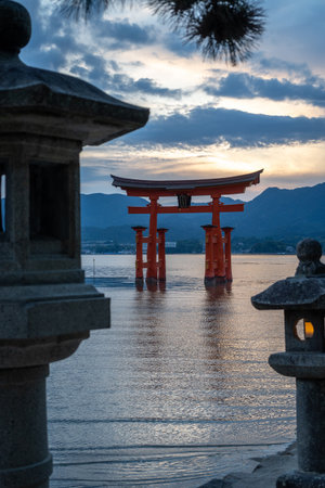 The famous floating torii gate of Itsukushima Shrine stands in calm waters at sunset, framed by stone lanterns and distant mountainsの写真素材