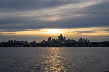 The Sydney skyline is silhouetted against a dramatic sunset, with golden hues reflecting on the harbor. The clouds add depth and contrast to the cityscapeの写真素材
