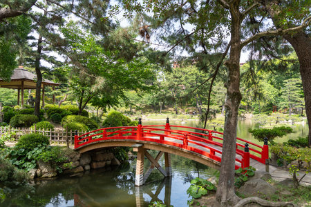 A vivid red arched bridge spans a pond in the peaceful Shukkei-en Japanese garden located in Hiroshima, surrounded by trees and greeneryの写真素材