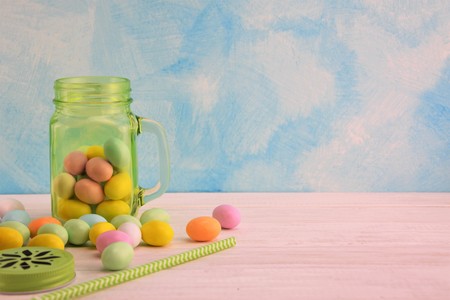 Green transparent glass jar with colored Easter eggs and straw on the white wooden table and...