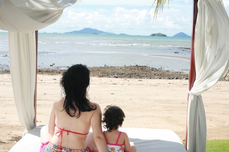 young lovely mother and her little daughter on tropical white bed close to the beach in the glow of the evening sunの写真素材