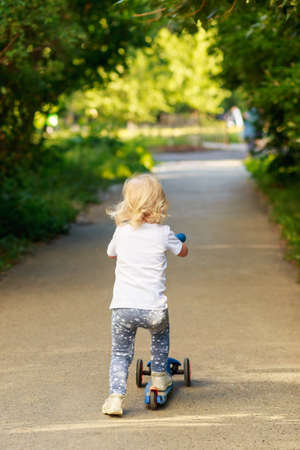 Happy beautiful girl is riding in the summer along the street on the three-wheeled scooter on a...