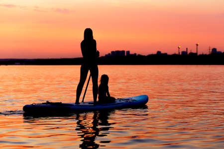 Silhouettes of woman and baby paddling on paddle board at sunset. Water sport near the beach on...