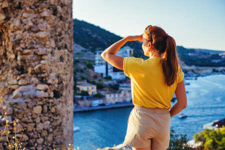 A girl in a yellow shirt admires the sunset against the background of a Balaklava bay and the ruins of an ancient Cembalo fortressの写真素材