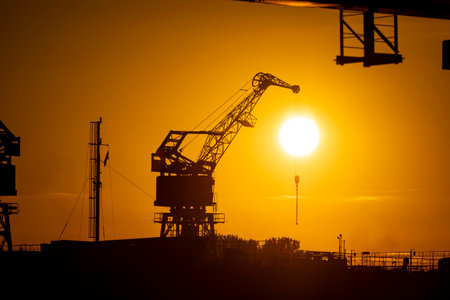 Beautiful metal crane silhouettes against the setting sun disc in harbour. Industrial cityscape of Riga, Latvia.の写真素材