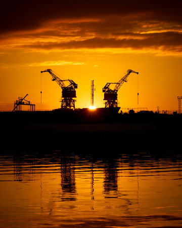 Beautiful metal crane silhouettes against the setting sun disc in harbour. Industrial cityscape of Riga, Latvia.の写真素材