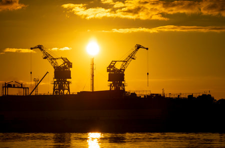 Beautiful metal crane silhouettes against the setting sun disc in harbour. Industrial cityscape of Riga, Latvia.の写真素材