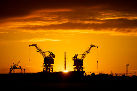Beautiful metal crane silhouettes against the setting sun disc in harbour. Industrial cityscape of Riga, Latvia.の写真素材