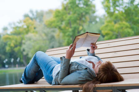 Young woman reading a book lying on the bench in the park