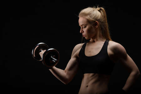 Young slim strong muscular woman posing in studio with dumbbell on black backgroundの写真素材