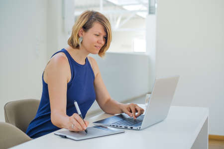 Young white female designer working on a laptop using a graphic pen tablet in the office.