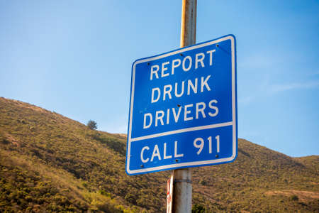 From below of blue sigh road with inscription Report drunk drives call 911 with green hillside and...