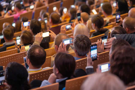 Business conference attendees sit and listen to lecturer, take pictures using smartphones, rear top viewの写真素材