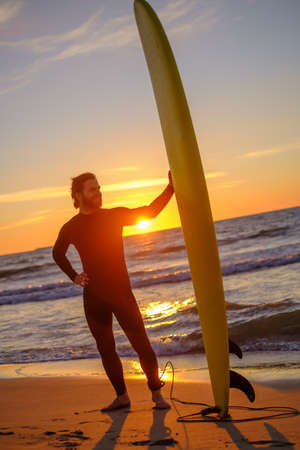 Full body bearded man with surfboard standing on sandy beach against waving sea and sundown sky on resortの写真素材