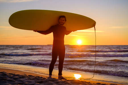 Full length happy bearded man in wetsuit carrying surfboard on shoulders while resting on sandy...