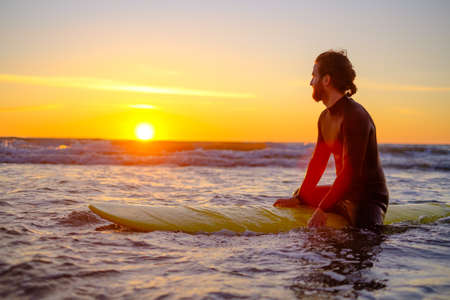 Side view of man in wetsuit sitting on surfboard and resting in sea while surfing with friend during...
