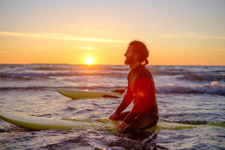 Side view of man in wetsuit sitting on surfboard and resting in sea while surfing with friend during sunsetの写真素材
