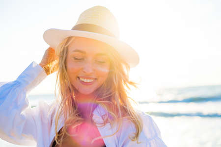 Positive female wearing sunhat and bikini smiling and enjoying holiday while looking away on background of waving seaの写真素材
