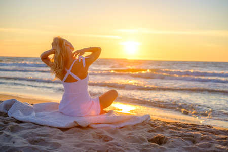 Back view of relaxed female traveler in summer dress sitting on blanket on sandy shore and admiring picturesque sunset over waving seaの写真素材
