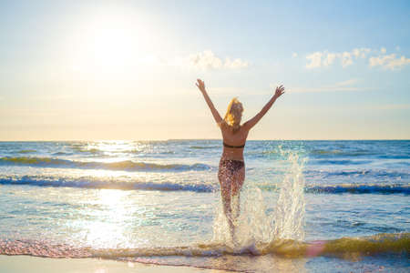 Back view of anonymous female in bikini running to sea water and enjoying sunny weather with raised armsの写真素材