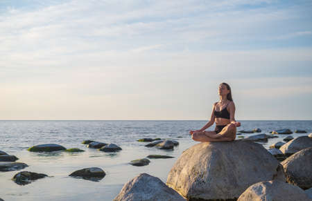 Young female sitting on boulder in Lotus pose and meditating against cloudy sky while doing yoga on seashoreの写真素材