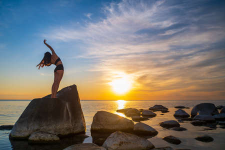 Back view of unrecognizable graceful woman bending aside with raised arms while dancing on stone near sea during sundownの写真素材