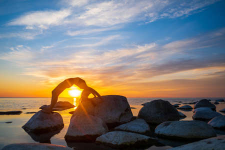 Side view of unrecognizable woman doing Wheel pose on boulders near sea against vibrant sunset sky