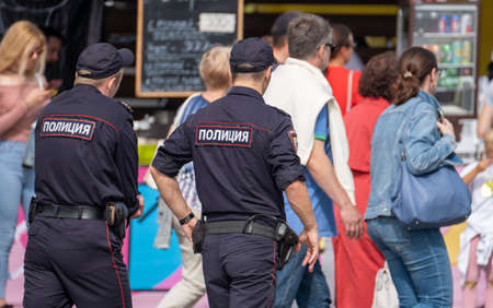 Back view of two men in police uniform walking on pavement and patrolling fairground in city