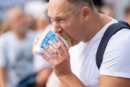 Kaliningrad, Russia - August 12, 2020: Plump mature male biting delicious takeaway sandwich and looking away on blurred background of crowd on city streetのeditorial素材