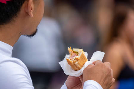 Unrecognizable guy holding tasty hot dog wrapped in paper while standing in crowd on city street