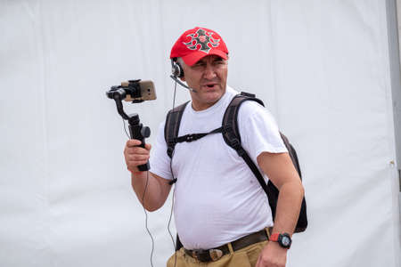 Kaliningrad, Russia - August 12, 2020: Side view of middle aged male with backpack walking on street and recording video on summer day in city using smartphone and 3 axis stabilizerのeditorial素材