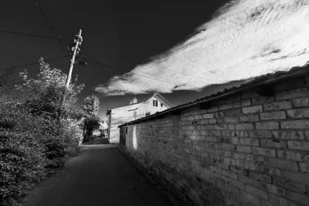 Black and white of empty narrow pathway leading between weathered brick fence and bushes under cloudy sky in summer day in countrysideの写真素材