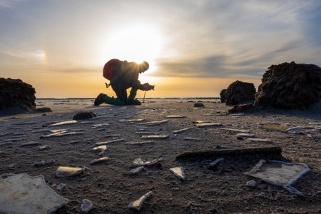 Silhouette of a volunteer actively picking up trash on a littered beach against a vivid sunset...