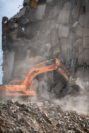An orange excavator is actively demolishing a large building, creating a cloud of dust and debris in...