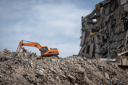 An excavator at a demolition site with a large pile of rubble and debris, next to a partially...