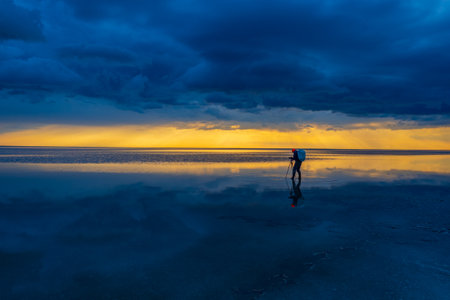 A lone traveler with a backpack walks across calm, reflective water under dramatic skies at sunset. Adventurous, peaceful, and inspiring.の写真素材