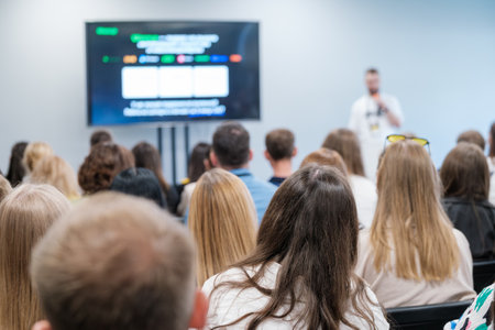 Diverse audience attentively listening to a speaker during a business presentation in a modern conference room, with a large screen displaying information.の写真素材