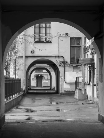 A black and white photograph of an urban courtyard with multiple archways, wet ground, and old...