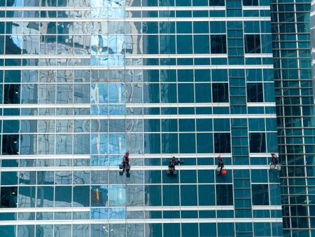 Four window cleaners suspended on ropes, cleaning a modern glass buildings windows, reflecting an urban cityscape. High-rise maintenance in progress.の写真素材