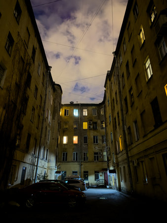Dark urban courtyard scene at night with cars parked and illuminated windows. The sky is cloudy and...
