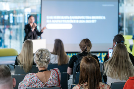 Attendees engaged in a corporate business presentation, listening to a speaker on stage in a modern...