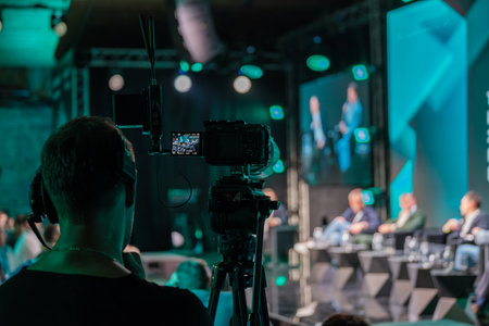 Close-up of professional video camera capturing speakers on stage during business conference. Focus on technology and communication in live event filming.の写真素材